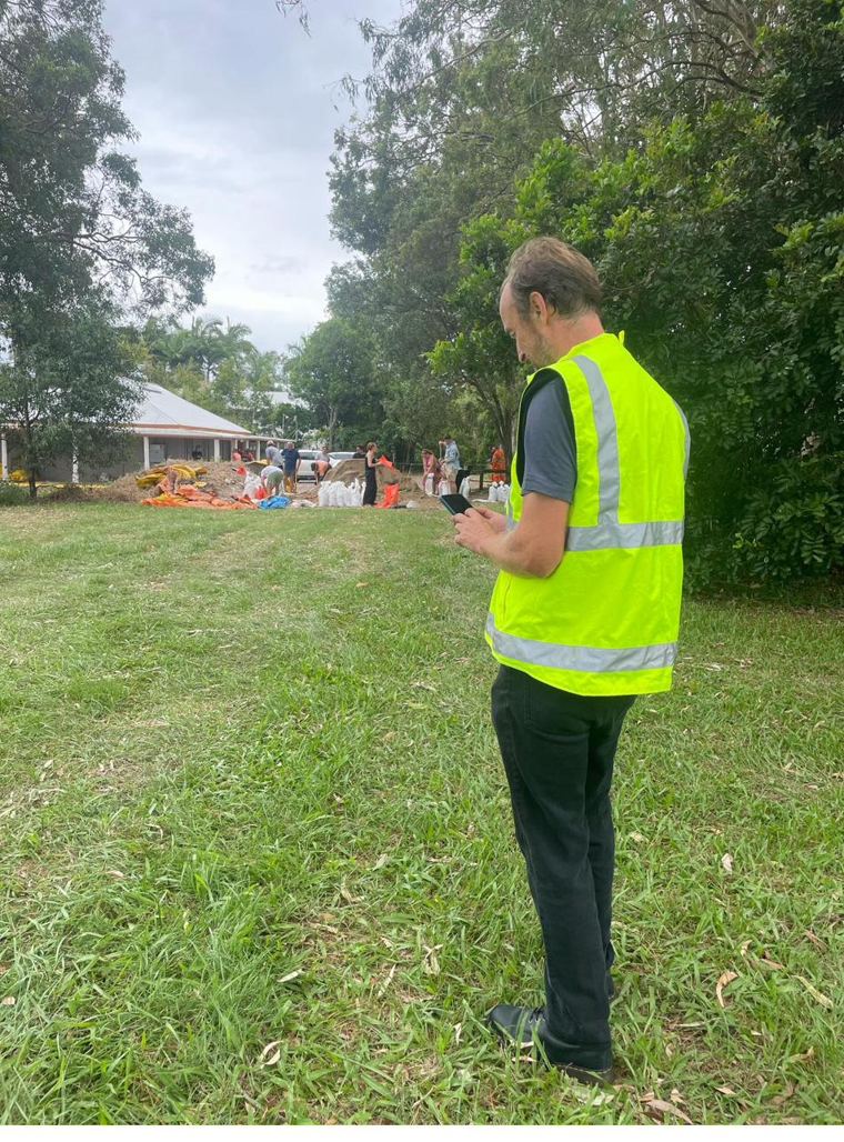 Leif Petersen in a high-visibility vest checking his phone while volunteers fill sandbags on a grassy lot